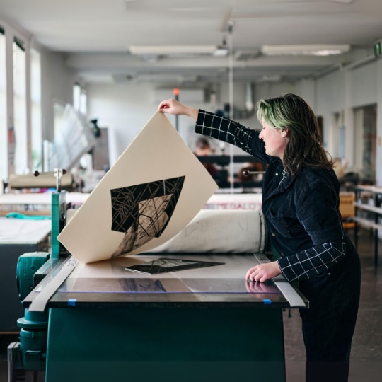 Photo of Bea Buckland-Willis in a print room, they have a green fringe and brown shoulder length hair, they are pulling up a print from a table and it is a geometrical shape, with dark black and white and grey shapes and lines